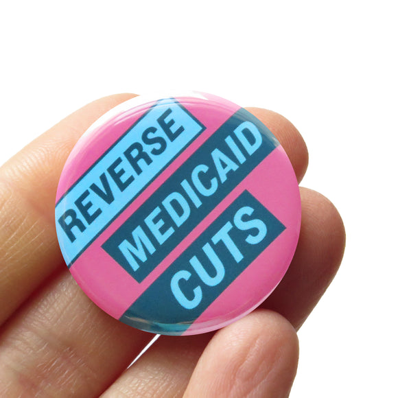 A round pink pinback button with light aqua text and dark teal boxes that reads REVERSE MEDICAID CUTS. The buttons held in a hand.