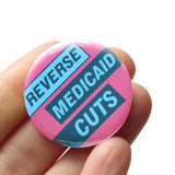 A round pink pinback button with light aqua text and dark teal boxes that reads REVERSE MEDICAID CUTS. The buttons held in a hand.