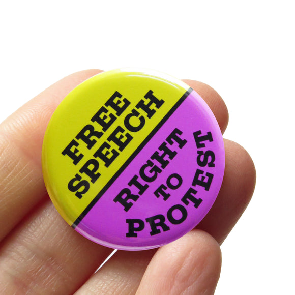 A round purple & lime green pinback button split down the middle. The green top half reads FREE SPEECH, the blue lower half reads RIGHT TO PROTEST. Buton is held in a hand.