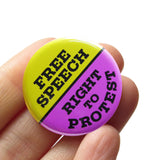 A round purple & lime green pinback button split down the middle. The green top half reads FREE SPEECH, the blue lower half reads RIGHT TO PROTEST. Buton is held in a hand.