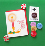 A 3-color risograph holiday card that features a red candle with a round yellow glow, dripping wax, in a classic gold holder with a spring of green holly berries. The red text reads Warmest Wishes in these Dark Times. A red envelope is with the card, and they sit on a green background, with a number of colorful holiday pinback buttons that read: Gay Apparel, Latke Lover, Egg Nog Fog, Naughty/Nice, Hot Cocoa Sipper, and some red & white peppermint buttons that look like the candy.