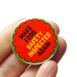 Round  & orange 1.25"  pinback button that reads MAKE FOOD SAFETY INSPECTED AGAIN in black and yellow text.  Badge is held in a hand.