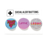 Three round pinback buttons in various vintage 1980's styles on a SOCIAL ALERT BUTTONS backing card. Buttons Left to Right: A blue button with a magenta triangle and gold text that reads POWER DYKE, A pink ombre button that reads SAPPHIC in a thin magenta pink font & a white button with tiny Memphis style black squiggles that reads LESBO in pink text with a Yellow drop shadow.