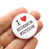 Round white pinback button that reads I LOVE SCIENCE FICTION. "Love" is represented by a red heart. Button is held in a hand.
