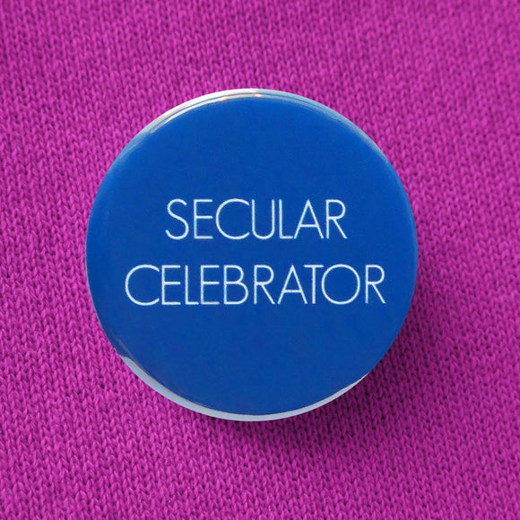 Bold bright blue round pinback button that reads SECULAR CELEBRATOR in thin white sans serif text.  Button is pinned to a raspberry sweatshirt.