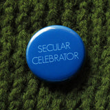 Bold bright blue round pinback button that reads SECULAR CELEBRATOR in thin white sans serif text.  Button is pinned to a green sweater