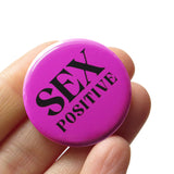 Round medium pink pinback button that reads SEX POSITIVE in black text, two lines.  Button is held in a hand.