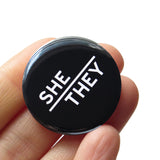 ound pinback button with black background which reads SHE/THEY  on an angle in white text, in a san serif font. Badge is held in a hand.