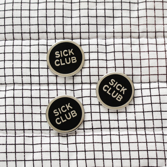 Round enamel pin that says SICK CLUB. Silver text and outline on a black background.