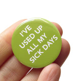 a round moss-lime green pinback button that reads I've Used Up All My Sick Days in white text. Button is held in a hand.