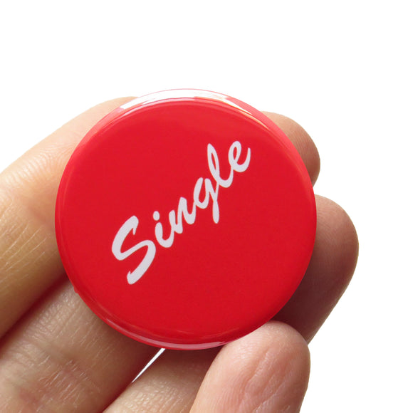 Round red pinback button that reads SINGLE in a white script font. Badge is held in a hand.