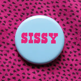 Round pinback button with a pale blue-teal background which reads SISSY in a bright pink chunky text.  BAdge is pinned to a vintage hot pink 80's shirt with tiny black triangles.