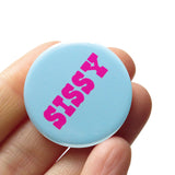 Round pinback button with a pale blue-teal background which reads SISSY in a bright pink chunky text.  Badge is held in a hand.