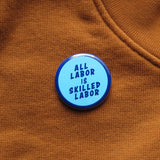 Round light blue pinback button with a dark blue outline & dark blue text that reads ALL LABOR IS SKILLED LABOR. The button is on a rich warm caramel colored sweatshirt.