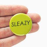 ound lime tree pinback button that reads SLEAZY in a think black text. Button held in a hand, by fingers.