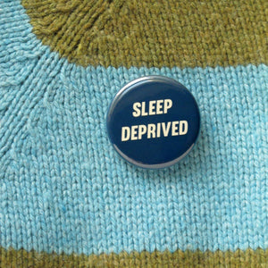 Round navy blue pinback button that reads SLEEP DEPRIVED in cream slanted text. Pinned to a light blue & olive green sweater.