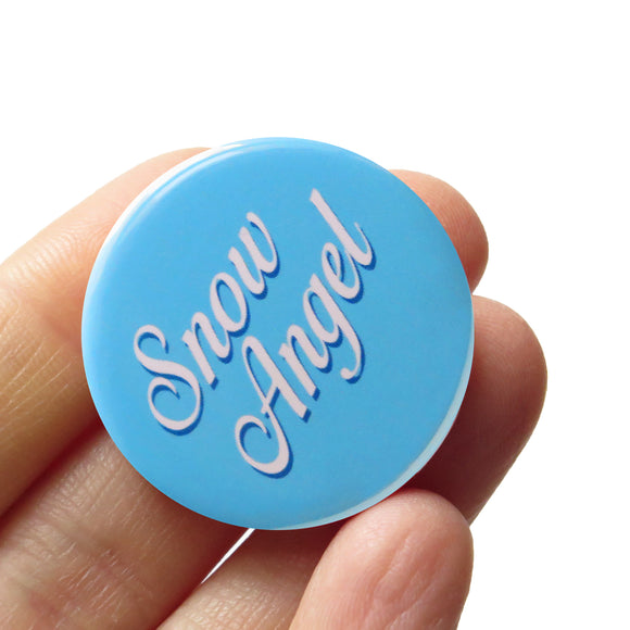 Light blue round pinback button that reads SNOW ANGLE in a white script font, which dark blue drop shadow, held at an angle, in hand