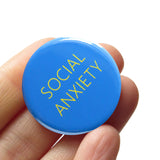 Round vibrant blue 1.25" pinback button with very thin yellow text reading SOCIAL ANXIETY. Button is held in a hand.