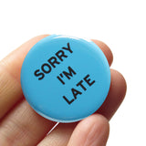 Round medium blue pinback button that reads SORRY I'M LATE in black text. Badge is held in a hand.