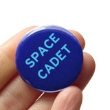 Round navy blue pinback button with vivid light blue text reading SPACE CADET. Badge is held in a hand.
