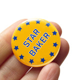 Round rich yellow pinback button that reads STAR BAKER in a blue san serif font with thin white outline, and border for blue stars. Badge is held in a hand.