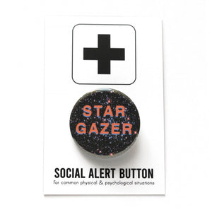 Round pinback button that reads, STAR GAZER. Salmon pink text with a light purple drop shadow, set on a background of outer space, full of stars. The button is pinned to a Social Alert Button backing card.