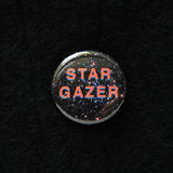 Round pinback button that reads, STAR GAZER. Salmon pink text with a light purple drop shadow, set on a background of outer space, full of stars. The button is pinned to a black wooly fabric.