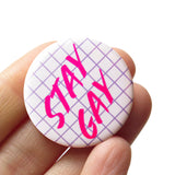 Round white pinback button with a lavender grid pattern reads STAY GAY in hot pink text. Button is being held by a white hand.