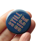 Dark blue round pinback button with hollowed neon yellow & pink text that reads STILL SICK. Button is held in hand.
