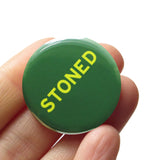 Round pinback button that says STONED. Light green text on a dark green background. Button is held in a hand.