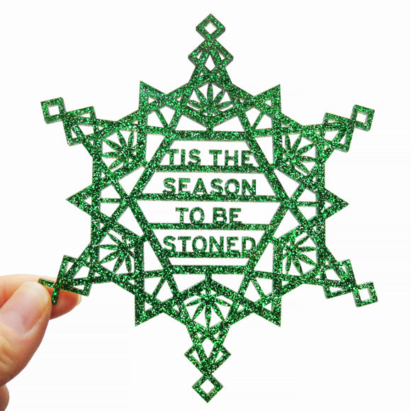 Snowflake shaped green glitter acrylic ornament, which in the center of the snowflake reads in san serif text on four lines: Tis The Season To Be Stoned. The cannabis snowflake is about 5 inches in size and is held in the lower left-hand corner by a thumb and forefinger