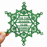 Snowflake shaped green glitter acrylic ornament, which in the center of the snowflake reads in san serif text on four lines: Tis The Season To Be Stoned. The cannabis snowflake is about 5 inches in size and is held in the lower left-hand corner by a thumb and forefinger