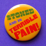 A round yellow-orange pinback button that reads STONED AND IN TERRIBLE PAIN! in green. purple & red text.  Button is on a purple background.
