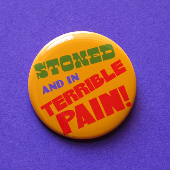 A round yellow-orange pinback button that reads STONED AND IN TERRIBLE PAIN! in green. purple & red text.  Button is on a purple background.