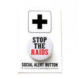 A round white pinback button that reads STOP THE RAIDS in black & red text. Button is on a Social Alert Button backing card.