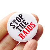 A round white pinback button that reads STOP THE RAIDS in black & red text. Button is held in a hand.