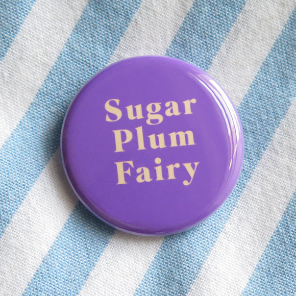 Shiny round purple pinback button that reads SUGAR PLUM FAIRY in rich cream text. On light blue & white stripped cotton shirt.