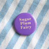 Shiny round purple pinback button that reads SUGAR PLUM FAIRY in rich cream text. On light blue & white stripped cotton shirt.