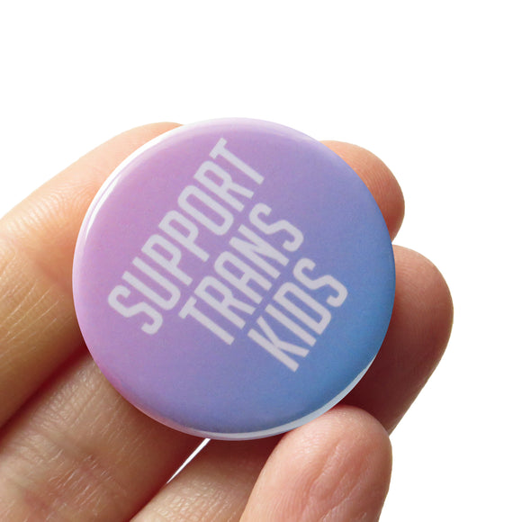 Round light pink & blue pinback button that reads SUPPORT TRANS KIDS. Button is an ombre gradient of light pink to light blue  with white san serif text. Button is held in a hand.