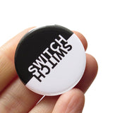 Round Pinback button that is black on the top half, and white on the bottom half.  The word SWITCH is in the inverse color on each side. Badge is held in a hand.