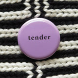 A round lavender 1.25" pinback button that reads tender in small, lower case black text. Button is pinned to a black and white chunky scarf.