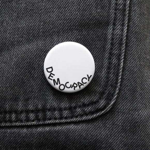 A round white pinback button that reads DEOMCRACY in black letters fallen to the bottom of the button.  Pinned on black denim.