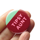 A round pinback button that looks like a tilted glass of wine. The bottom half is wine red burgundy with an angled top, mint green above. And inside the wine color it reads TIPSY AUNT in pink askew letters. Badge is held in a hand.