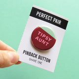 A round pinback button that look like red wine sloshing in a round cup with a mint green background. In the red white color it reads: TIPSY AUNT in pink. Backing card torn at perferation held by hand