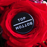 Round pinback button that says TOP and directly underneath it, upside down, its says BOTTOM.  White text on a black background.  The button is nestled inside a red rose