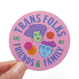 A round pink sticker that reads TRANS FOLKS ARE MY FRIENDS & FAMILY around the permitter of the sticker in periwinkle & green text, with two illustrated faces in the middle, one with minty skin, purple hair and pink lips. The other with blue skin, glasses and a wavy swoosh of magenta hair. Little pink, orange & yellow flowers are decorating the negative space around them. Sticker is held in the corner with a thumb and forefinger.