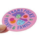 A round pink sticker that reads TRANS FOLKS ARE MY FRIENDS & FAMILY around the permitter of the sticker in periwinkle & green text, with two illustrated faces in the middle, one with minty skin, purple hair and pink lips. The other with blue skin, glasses and a wavy swoosh of magenta hair. Little pink, orange & yellow flowers are decorating the negative space around them. Sticker is held at an angle in the corner with a thumb and forefinger.
