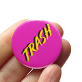 A round magenta pink pinback button that reads TRASH in a yellow font with a black drop shadow. Button is held in a hand.