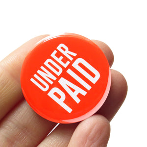 Bright round red-orange pinback button that reads UNDER PAID in white san serif text. Button held in a hand.