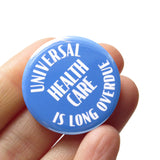 A round scrubs blue 1.25" pinback button that reads UNIVERSAL HEALTHCARE IS LONG OVERDUE in white text. Button is held in a hand.